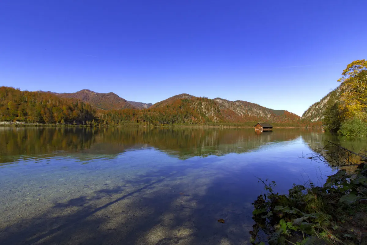 Ein klarer Bergsee zeigt die Reflexion der herbstlich gefärbten Bäume, während eine Holzhütte am rechten Ufer steht. Der Himmel ist strahlend blau.
