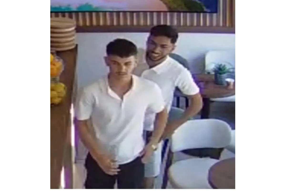 Zwei junge Männer in weißen Hemden stehen in einem Café. Einer hält eine Flasche, der andere lächelt.