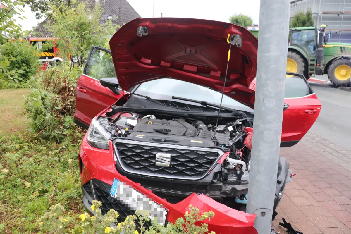Ein rotes SEAT-Fahrzeug ist frontal mit einem Laternenpfahl kollidiert. Der Motorraum ist offen und zeigt Schäden. Im Hintergrund ist ein Feuerwehrfahrzeug sichtbar.