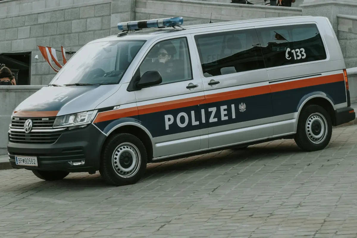 Ein österreichisches Polizeifahrzeug, ein silberfarbener VW-Bus mit blauen und roten Akzenten, steht auf einem gepflasterten Untergrund. Im Hintergrund sind Gebäudeteile und eine Person zu sehen.