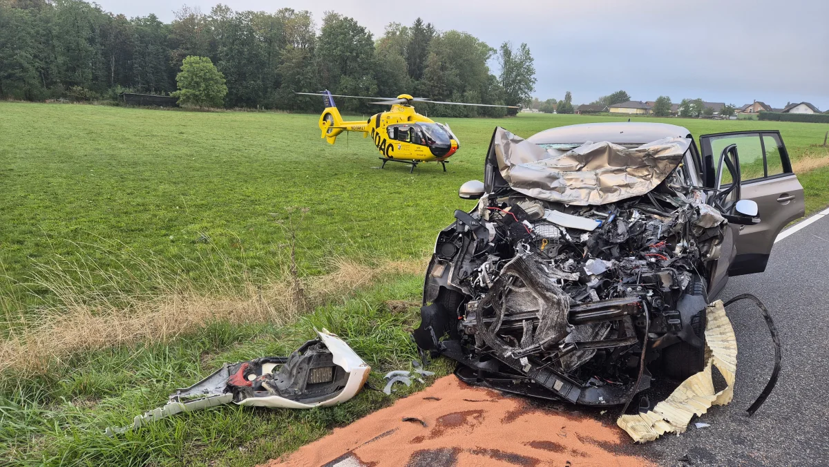 Ein stark zerstörtes Auto steht am Straßenrand einer Landstraße. Ein gelber Rettungshubschrauber befindet sich auf einem angrenzenden Feld.