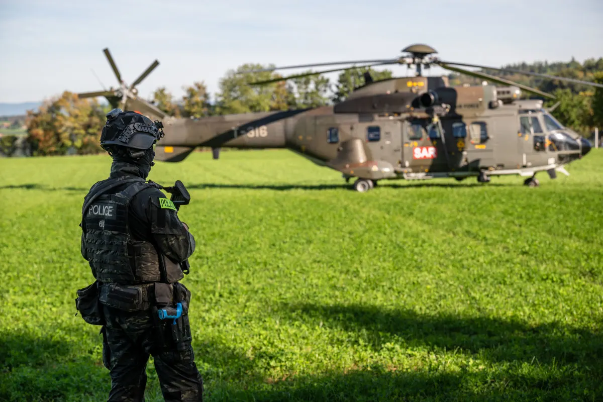 Ein Polizist in taktischer Ausrüstung steht im Vordergrund, während im Hintergrund ein SAR-Hubschrauber auf einem grünen Feld gelandet ist.