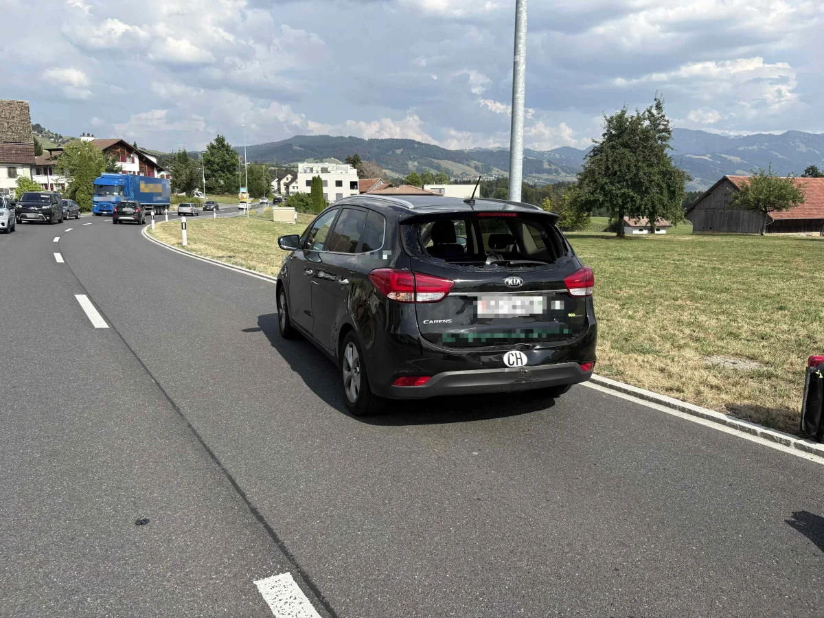 Ein schwarzes Auto steht mit beschädigtem Heck auf einer Landstraße. Im Hintergrund sind Häuser und eine hügelige Landschaft zu sehen.