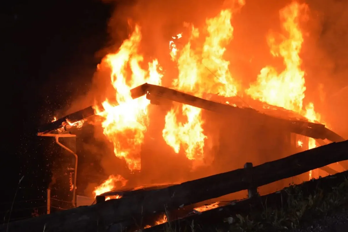 Ein Holzgebäude brennt nachts lichterloh, große Flammen schlagen aus dem Dachbereich. Der Himmel ist dunkel und von den leuchtenden Flammen kontrastiert.