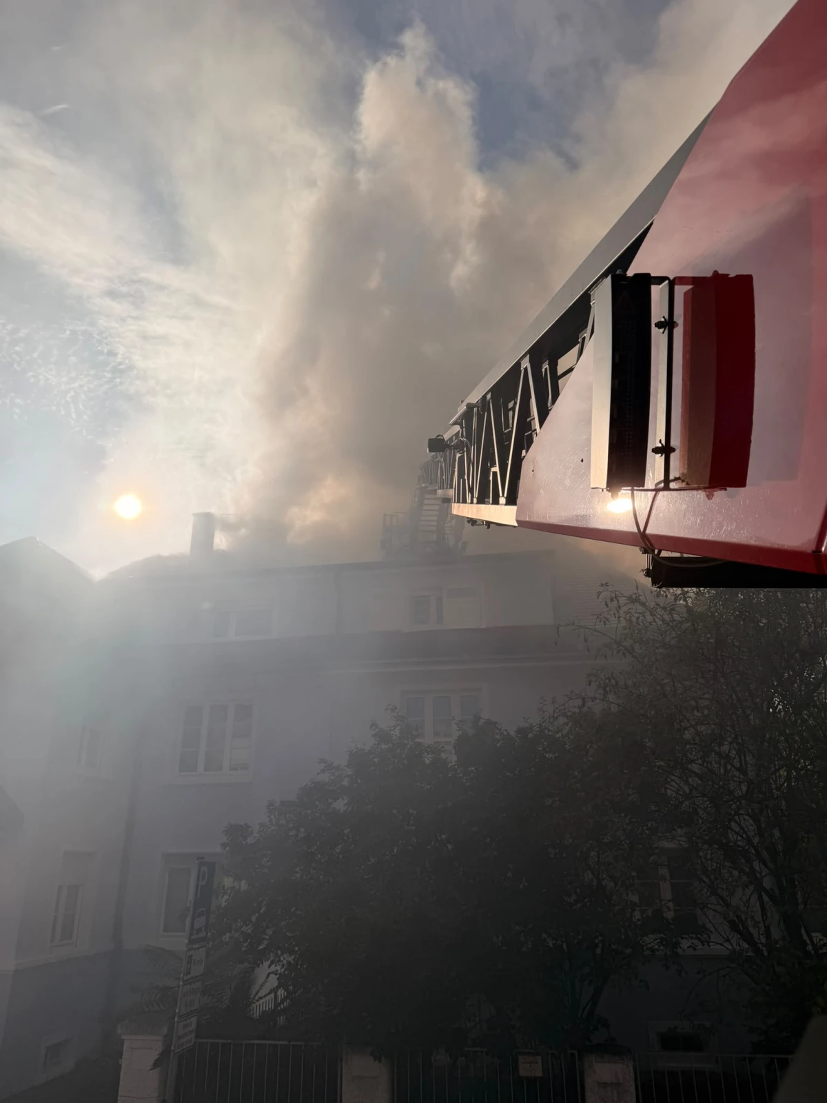 Eine Feuerwehrleiter ist vor einem Gebäude zu sehen, aus dessen oberen Etagen Rauch aufsteigt. Die Sonne scheint durch den Rauch.