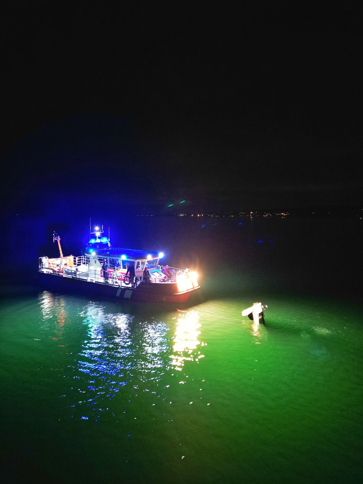 Ein Rettungsboot mit Blaulicht navigiert auf einem dunklen See. Im Wasser ist ein gesunkenes Wrack sichtbar.