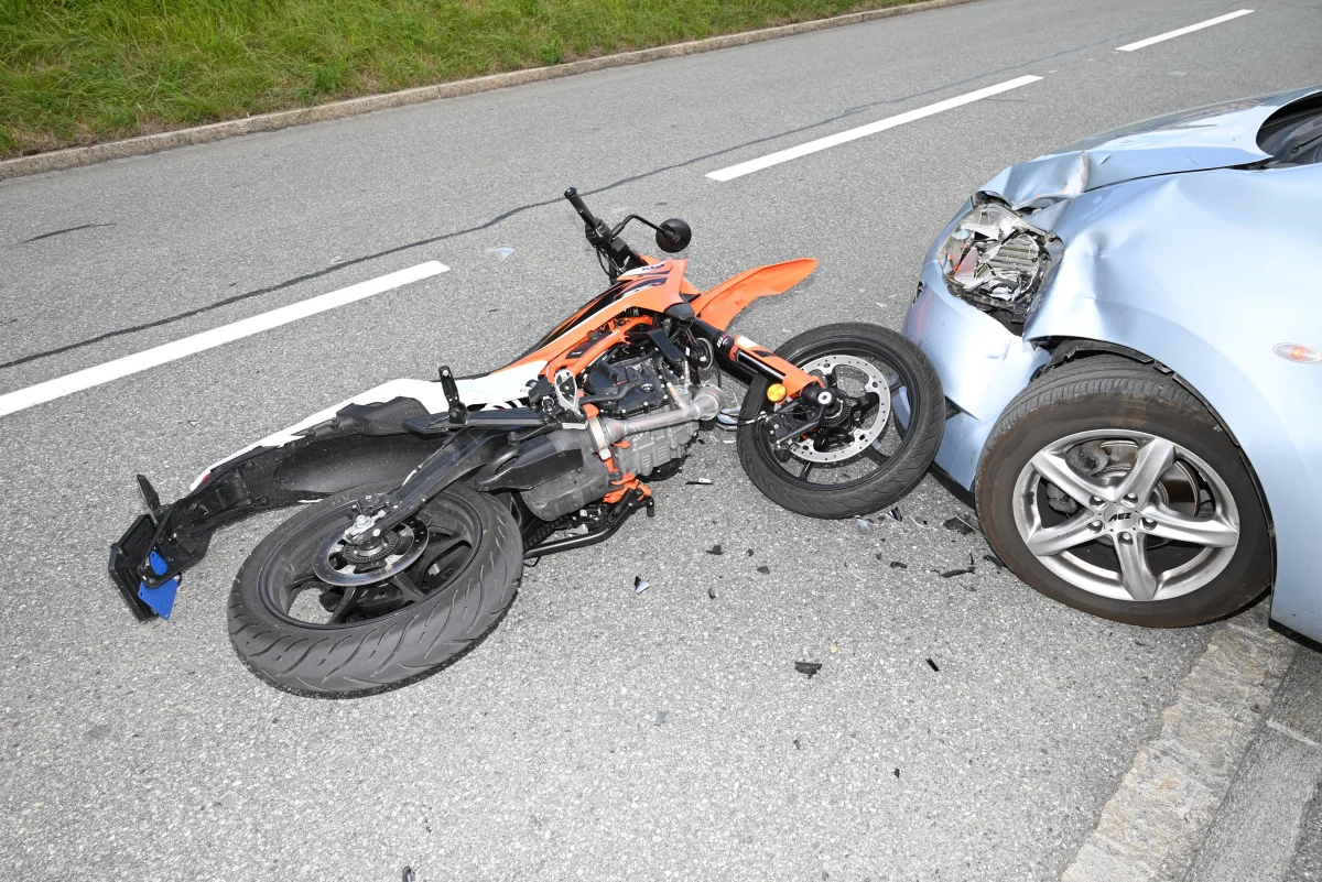 Ein orange-schwarzes Motorrad liegt auf einer Straße und kollidierte mit einem blauen PKW, dessen Front beschädigt ist. Einzelne Fahrzeugteile sind auf der Fahrbahn verstreut.