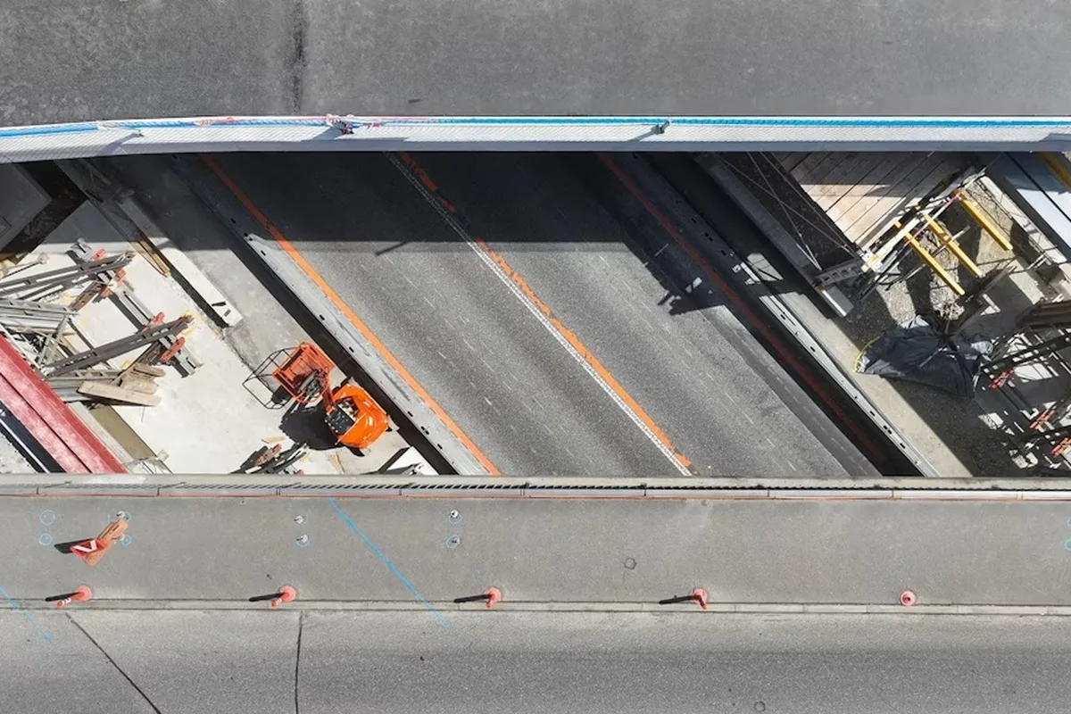 Ein Luftbild zeigt eine Baustelle mit einer orangefarbenen Baumaschine neben einer Straße, die durch Barrieren und Hütchen abgesperrt ist.