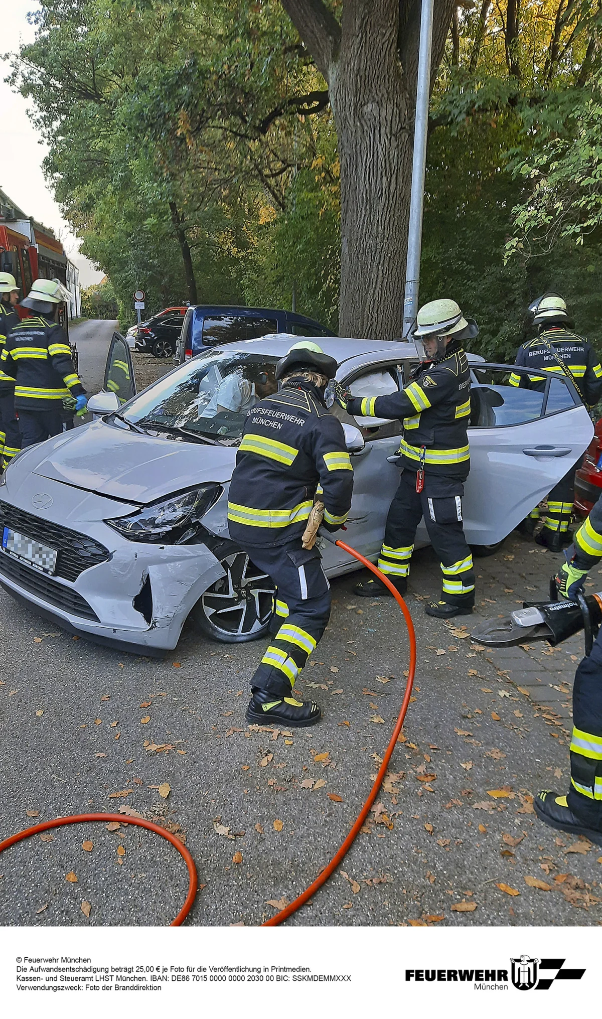 Mehrere Feuerwehrleute der Berufsfeuerwehr München retten eine Person aus einem beschädigten weißen Auto, das in einer Baumallee steht.