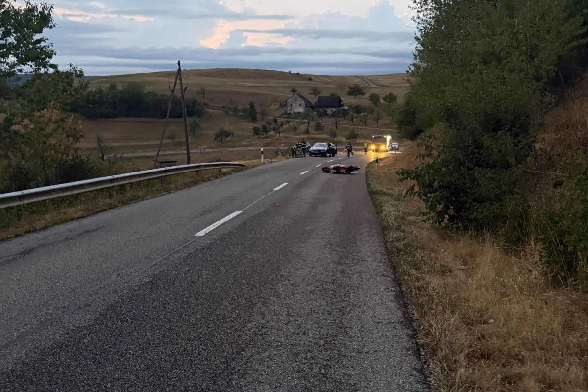 Ein verlassenes Motorrad liegt auf einer Landstraße, während Rettungskräfte im Hintergrund sichtbar sind. Die Szene findet bei Dämmerung statt, mit ländlicher Umgebung und einem Hügel im Hintergrund.
