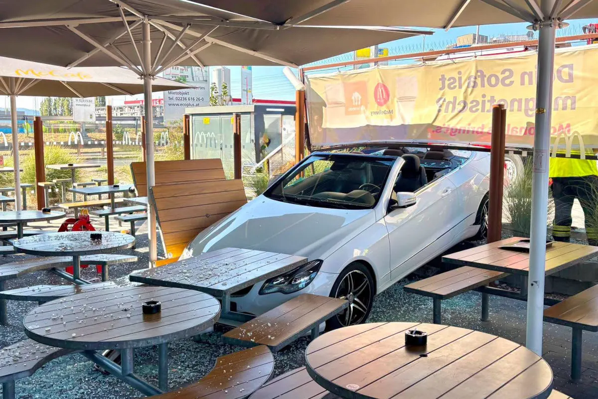 Ein weißes Cabrio steht in einem Außensitzbereich eines Cafés, umgeben von Tischen und umgekippten Bänken. Ein Arbeiter im gelben Anorak im Hintergrund.