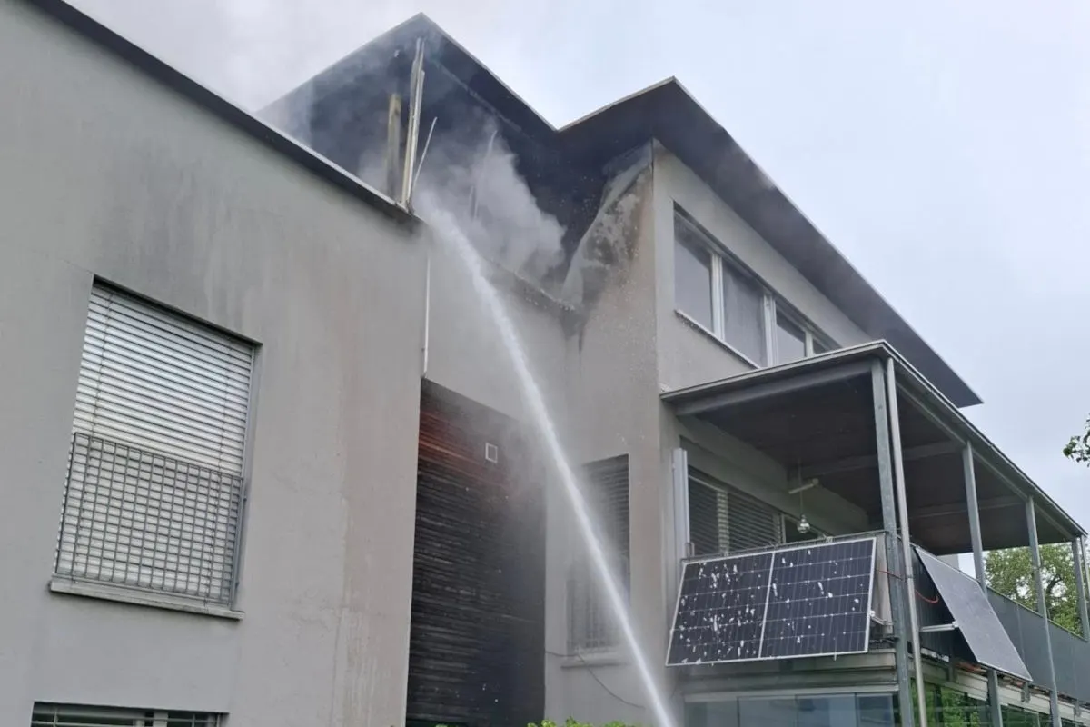 Ein Gebäude zeigt Rauch und Schäden am oberen Stockwerk, während ein starker Wasserstrahl von unten darauf gerichtet ist, um den Brand zu löschen. Solarpaneele sind an der Fassade sichtbar.