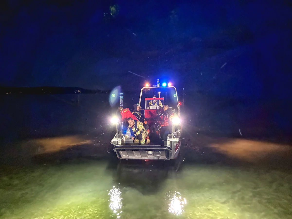 Ein Boot mit Einsatzkräften beleuchtet die Wasserfläche bei Dunkelheit. Lichter der Einsatzfahrzeuge sind sichtbar.