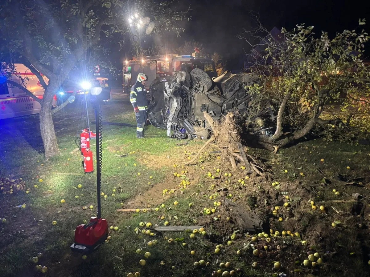 Ein umgestürztes Auto liegt neben einem entwurzelten Baum, umgeben von vielen gefallenen Äpfeln. Feuerwehrleute arbeiten an der Unfallstelle, die bei Nacht gut ausgeleuchtet ist.