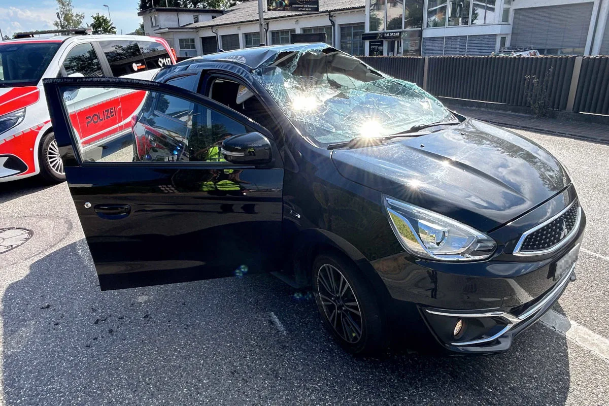 Ein schwarzes Auto mit stark beschädigter Windschutzscheibe steht mit offener Fahrertür auf der Straße. Daneben ist ein Polizeiwagen zu sehen.