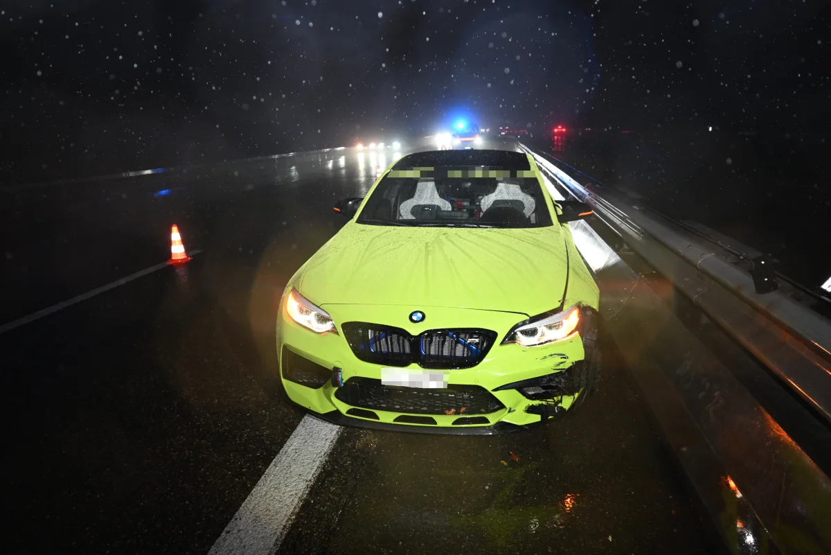 Ein grüner BMW mit sichtbaren Schäden steht auf einer nassen Straße. Im Hintergrund sind Polizeilichter und Absperrkegel zu sehen.