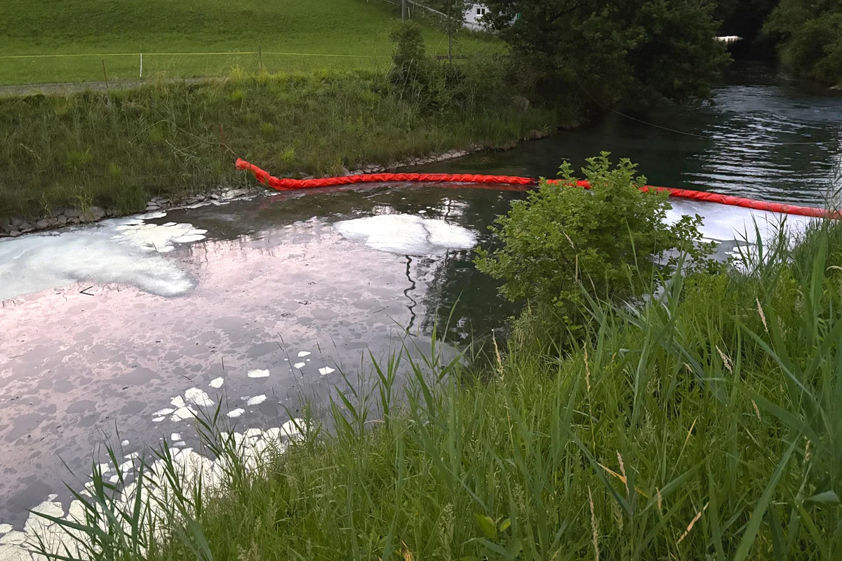 Eine rote Ölsperre verläuft quer über einen Fluss, der teilweise mit Schaum und Öl bedeckt ist. Im Hintergrund ist eine grasbewachsene Uferböschung zu sehen.