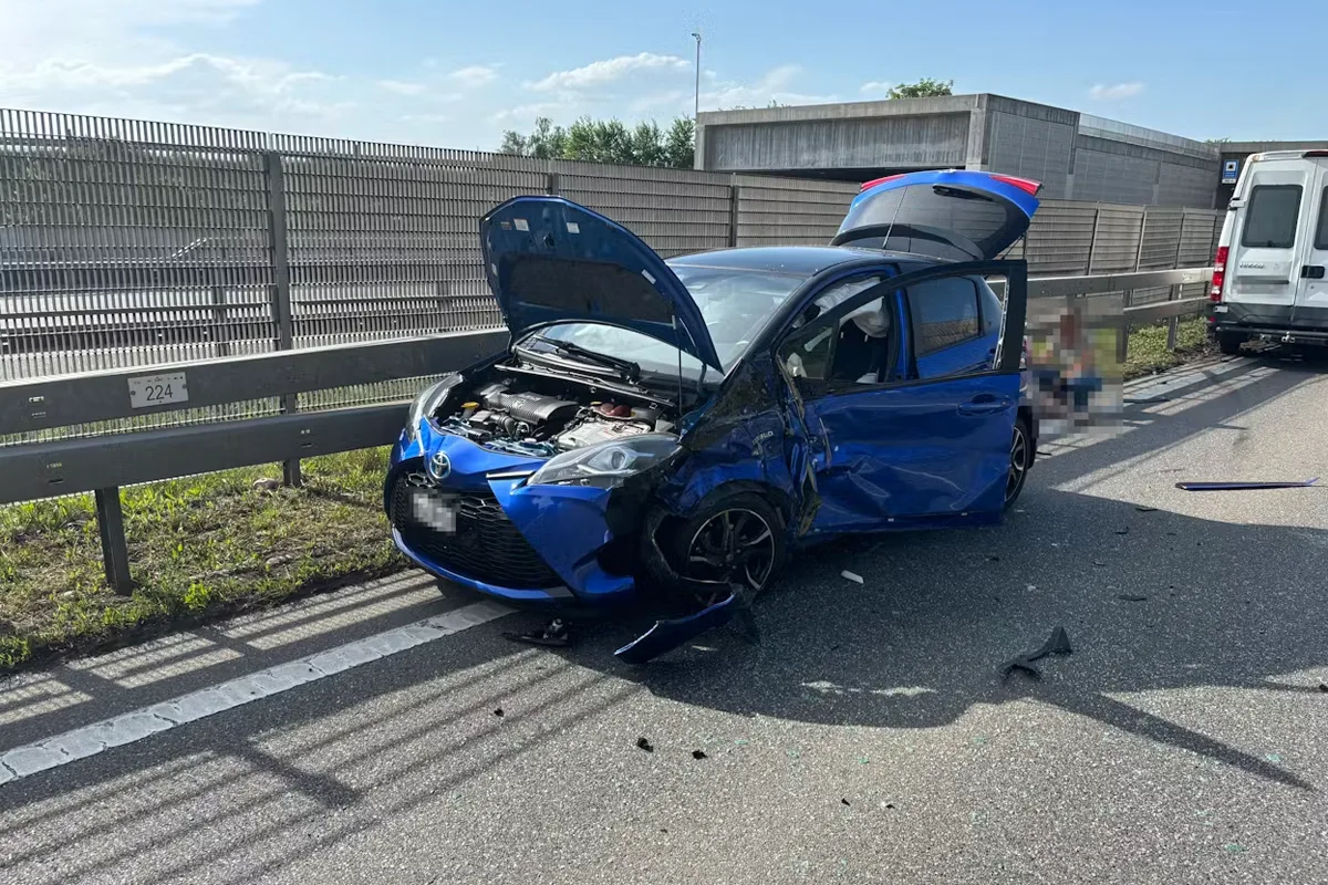 Ein blauer Kleinwagen steht mit geöffnetem Kofferraum und beschädigter Vorderseite am Rand einer Autobahn. Daneben erkennt man einen weißen Van.
