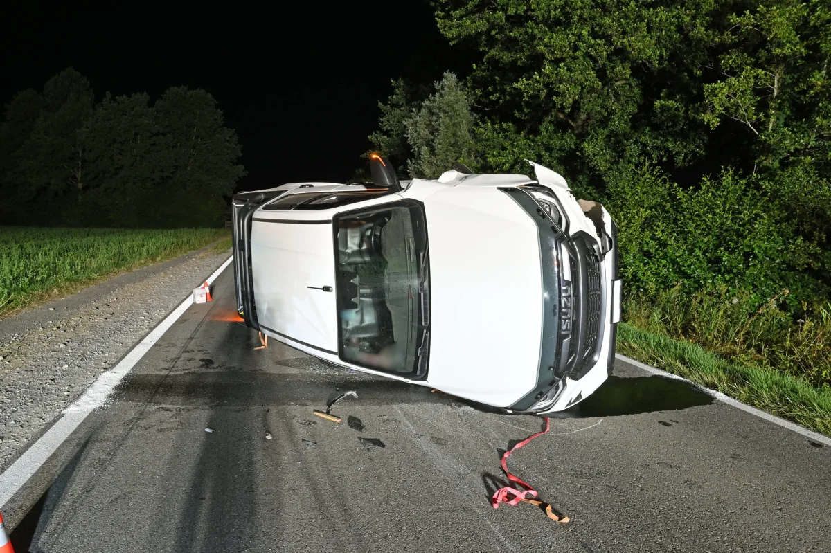 Ein weißes Auto liegt umgestürzt auf einer nächtlichen Landstraße, umgeben von Bäumen.