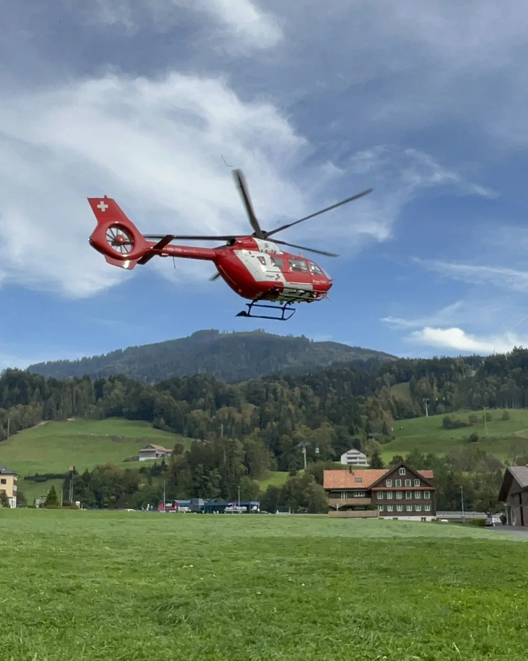 Ein roter Rettungshubschrauber ist im Flug über einer grünen Wiese mit bewaldeten Hügeln im Hintergrund. Das Wetter ist klar mit einigen Wolken.