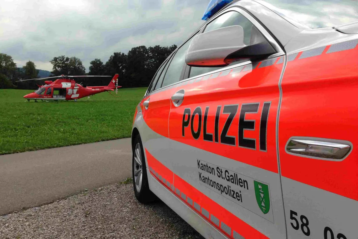 Ein Streifenwagen der Kantonspolizei St.Gallen steht am Rand einer Wiese, auf der ein roter Rettungshubschrauber gelandet ist. Der Himmel ist leicht bewölkt.