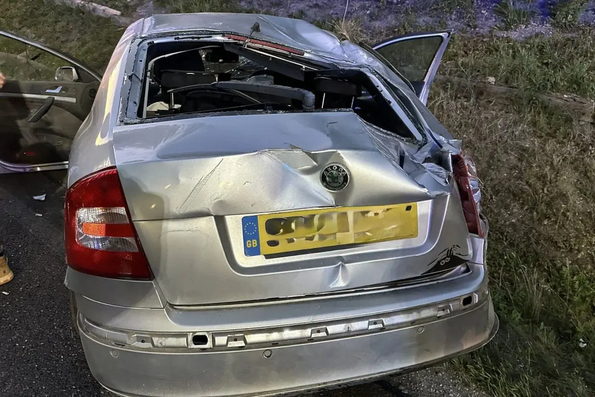 Ein beschädigter silberner Skoda steht mit stark eingedrücktem Heck und offener Hintertür auf einer Autobahn bei Nacht. Das Nummernschild zeigt GB.