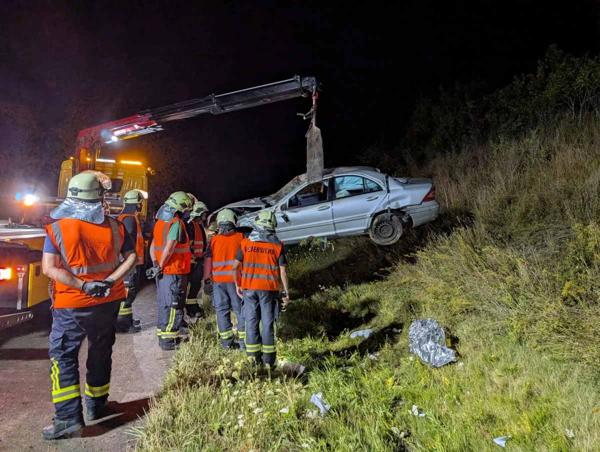 Mehrere Feuerwehrleute in orangefarbenen Westen beobachten, wie ein Auto bei Nacht mit einem Kran aus einem Graben geborgen wird.