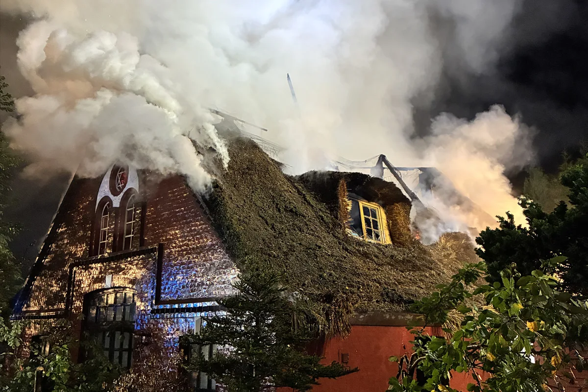 Ein Haus mit einem Reetdach brennt, dichter Rauch steigt auf. Das Gebäude ist von Flammen erfasst.