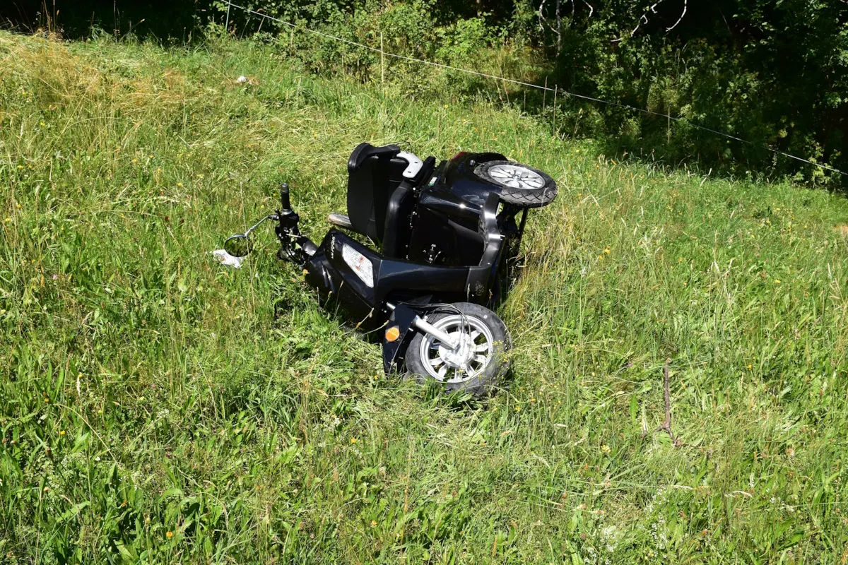 Ein schwarzer Roller liegt umgestürzt im hochgewachsenen Gras einer Wiese. Die Umgebung ist grün und ländlich.
