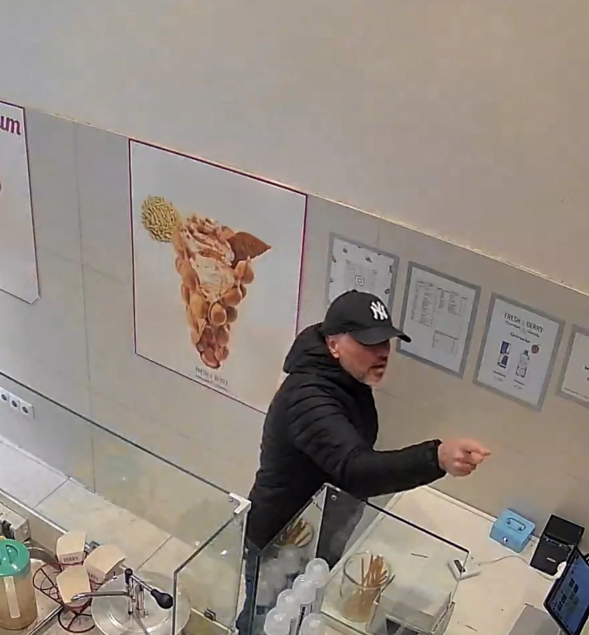 Ein Mann mit dunkler Jacke und Mütze gestikuliert an einem Tresen in einem Geschäft, aufgenommen von einer Überwachungskamera. An der Wand sind Plakate mit Eiscreme abgebildet.