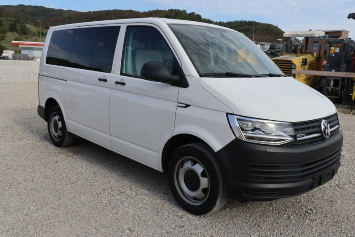 Das Bild zeigt einen weißen VW-Transporter, der auf einem Schotterplatz geparkt ist. Im Hintergrund sind Hügel und eine Tankstelle zu sehen.