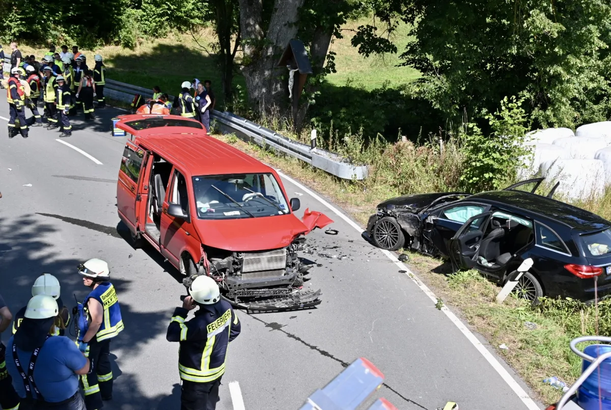 Ein roter Transporter ist frontal beschädigt und steht auf einer Straße. Rechts befindet sich ein schwarzer Pkw, ebenfalls beschädigt, am Straßenrand. Feuerwehrleute sind vor Ort.
