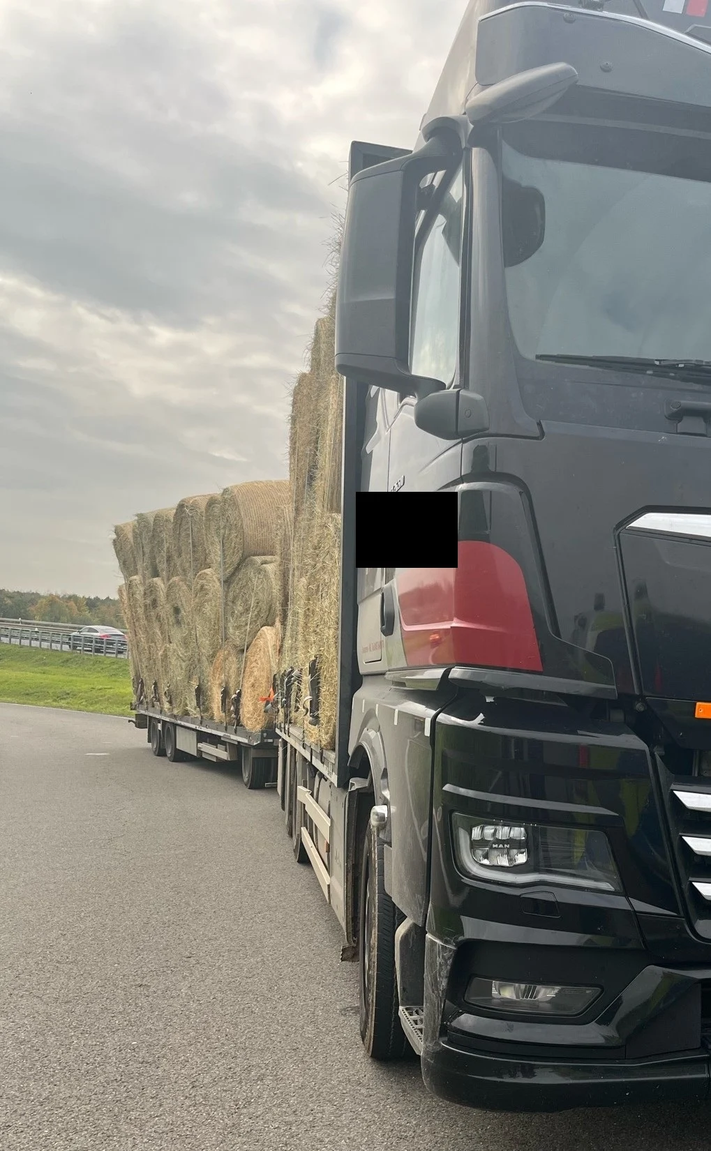 Ein schwarzer LKW ist auf einer Landstraße zu sehen, beladen mit vielen Heuballen auf einem Anhänger bei bewölktem Himmel.