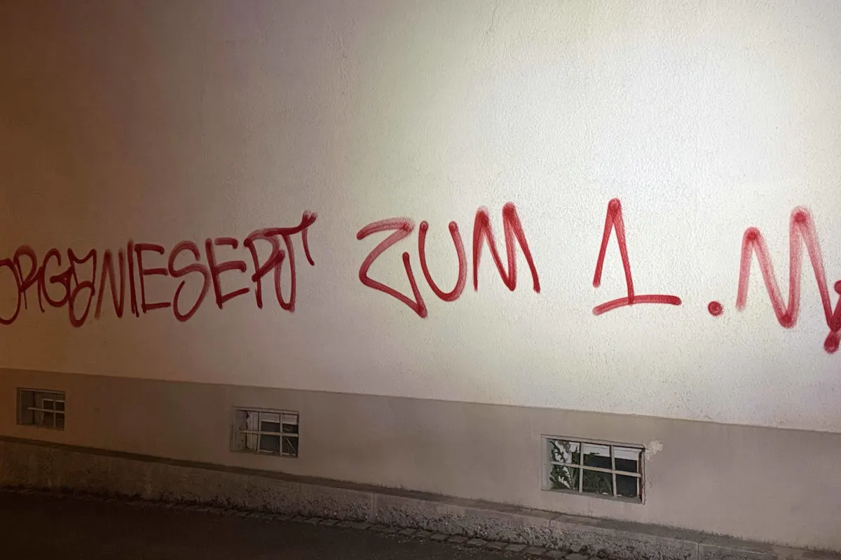 Eine helle Wand mit roter Graffiti-Schrift, die über den unteren Teil der Wand verläuft. Die Schrift ist in unregelmäßigen Abständen gesprüht.