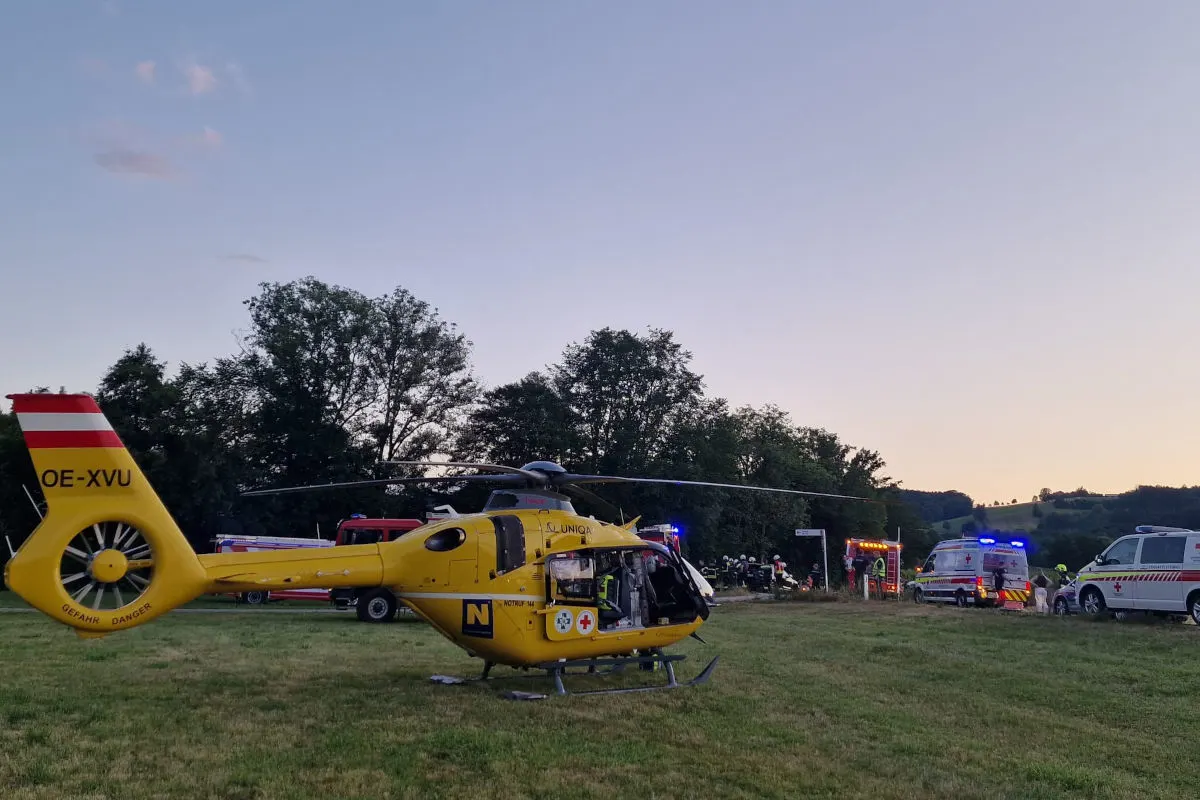 Ein gelber Rettungshubschrauber steht auf einer Wiese, umgeben von mehreren Einsatzfahrzeugen bei einbrechender Dämmerung. Im Hintergrund sind Bäume und Hügel sichtbar.