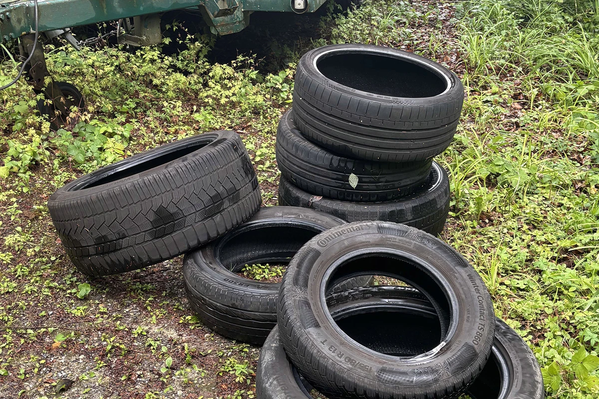 Auf einem Waldweg liegen mehrere gebrauchte Autoreifen übereinander gestapelt. Einige Reifen sind verunreinigt und die Umgebung ist dicht bewachsen.