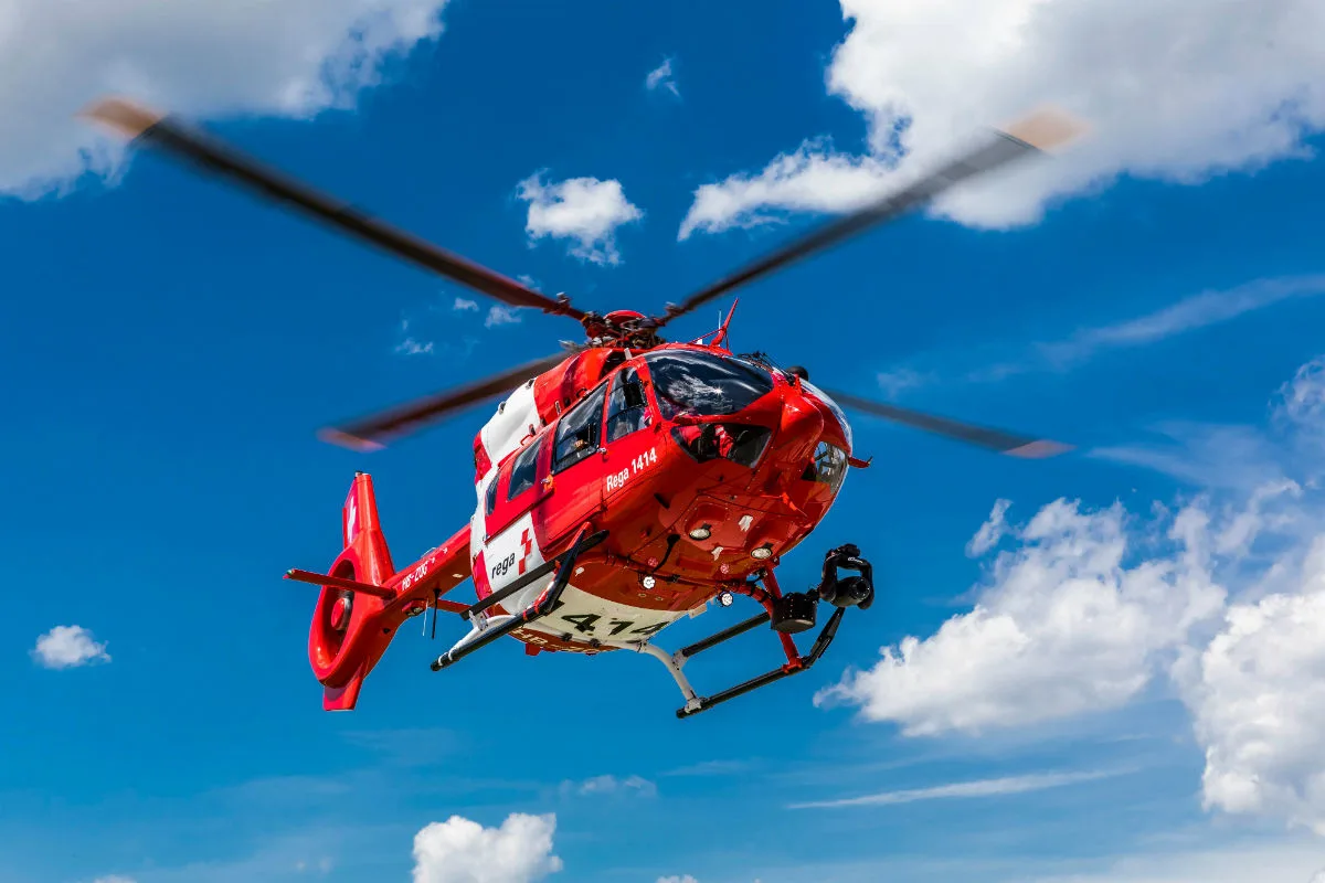 Ein roter Rettungshubschrauber ist in der Luft zu sehen, während er vor einem blauen Himmel mit wenigen Wolken fliegt. Der Hubschrauber trägt die Aufschrift 'Rega 1414'.