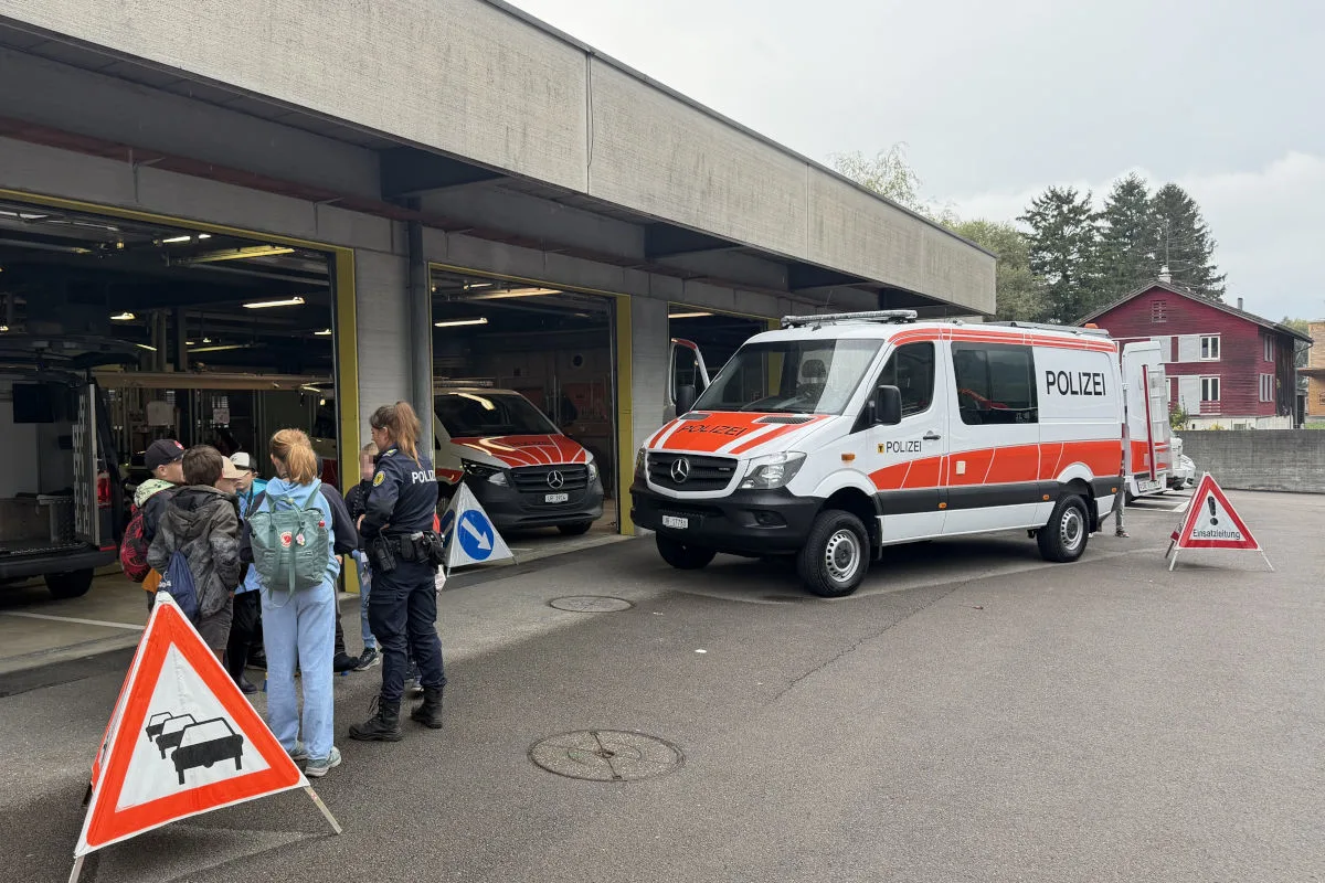 Ein Polizeifahrzeug steht vor einer offenen Halle, daneben sind eine Polizistin und mehrere Personen zu sehen.