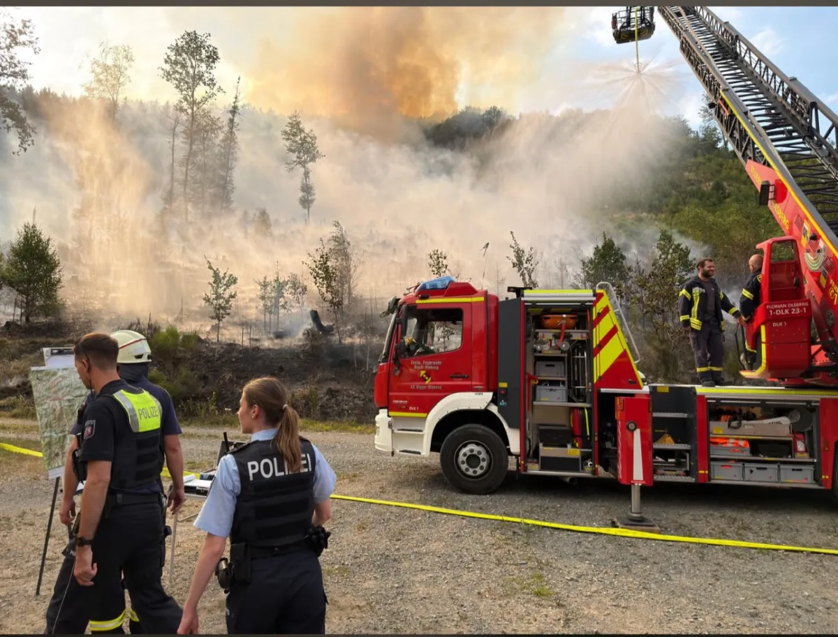 Ein Feuerwehrfahrzeug bekämpft eine Rauchentwicklung bei einem Waldbrand. Zwei Polizisten beobachten die Lage vor dem Fahrzeug.