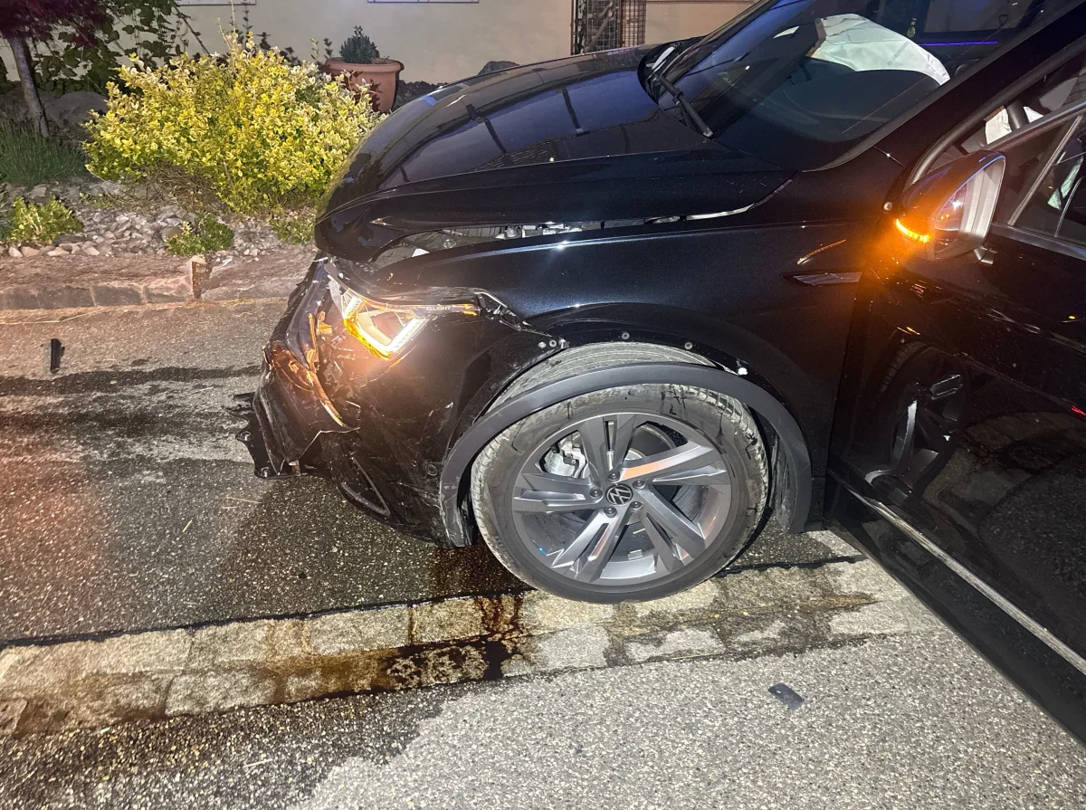 Ein schwarzer VW, dessen Vorderseite durch einen Zusammenstoß stark beschädigt ist, steht auf einer nassen Stadtstraße. Die Umgebung ist bei Nacht beleuchtet.