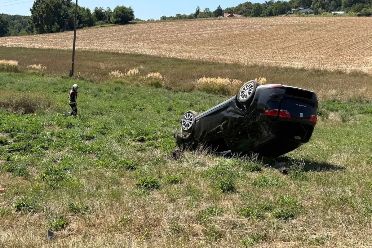 Ein schwarzes Auto liegt auf dem Dach in einem Feld. Ein Mensch in Schutzkleidung nähert sich dem Fahrzeug.