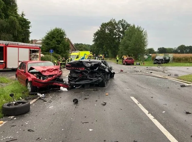 Zwei Autos mit schweren Frontschäden stehen auf einer Landstraße nach einer Kollision. Im Hintergrund sind Rettungsfahrzeuge und Einsatzkräfte sichtbar.
