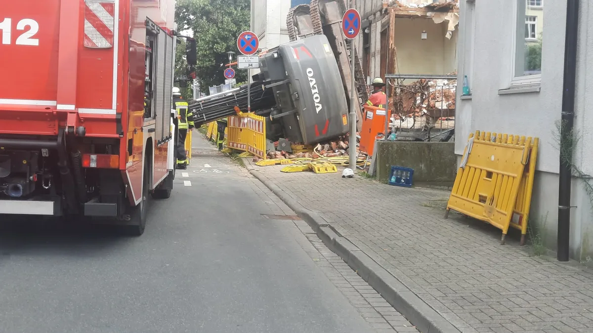Ein umgestürzter Bagger liegt neben einem beschädigten Gebäude. Feuerwehrkräfte sind vor Ort, um die Szene abzusichern.