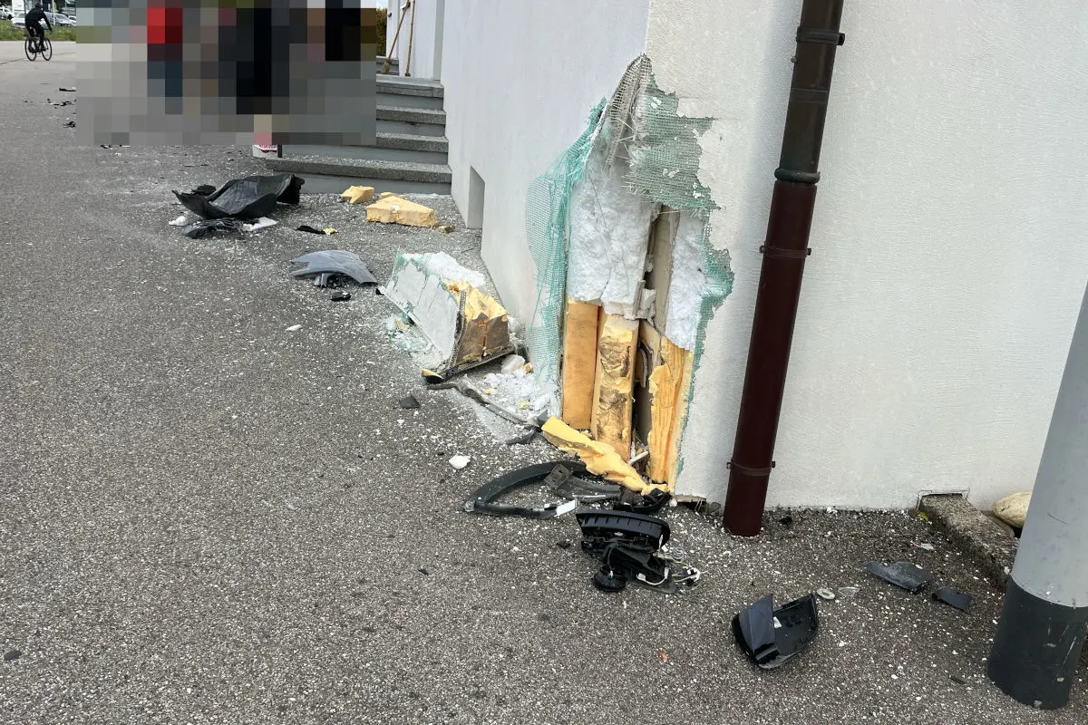 Die Wand eines Gebäudes zeigt deutliche Unfallspuren, mit abgeplatztem Putz und freiliegender Isolierung. Auf dem Gehweg liegen Trümmerteile verstreut.