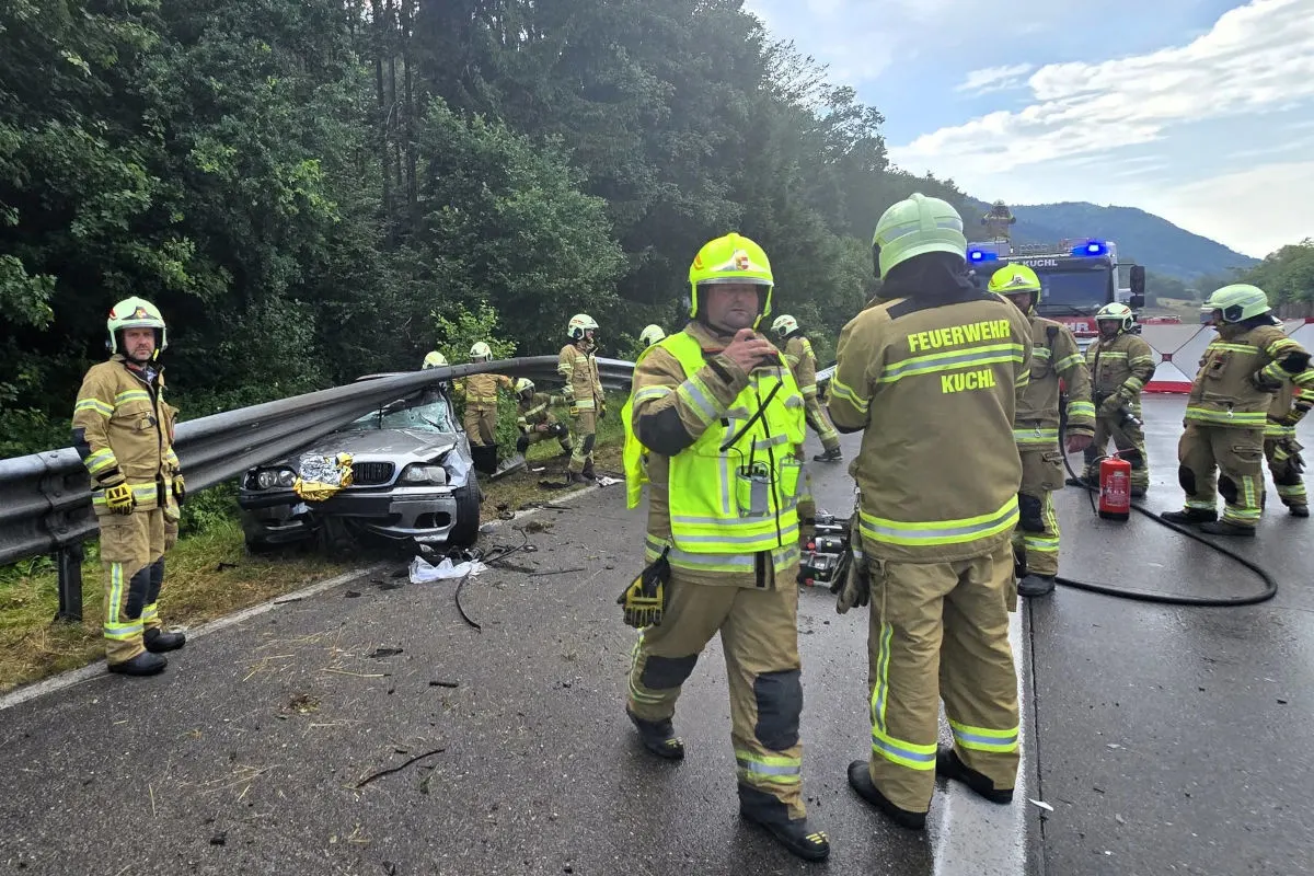 Ein Auto liegt verunfallt neben einer Leitplanke, umgeben von Feuerwehrleuten in Schutzanzügen auf einer Landstraße.