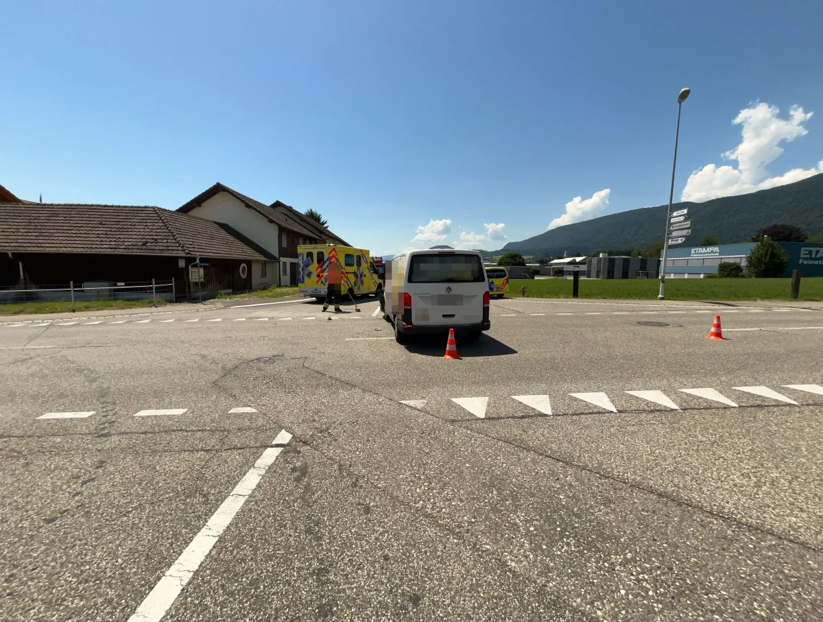 Ein gelber Krankenwagen mit roten Markierungen steht auf einer Landstraße, daneben ein weißer Transporter und zwei Verkehrskegel. Im Hintergrund sind Gebäude und bewaldete Hügel zu sehen, bei klarem Himmel und Sonnenschein.