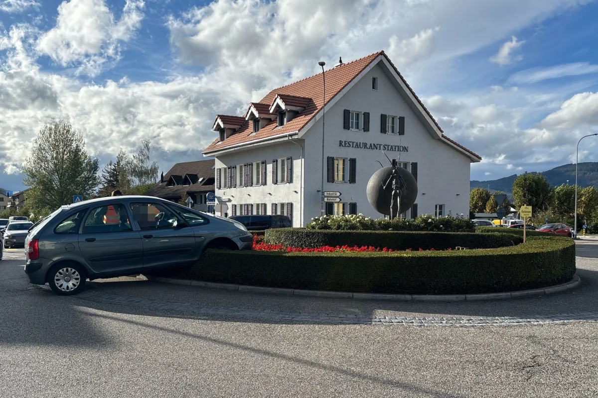 Ein graues Auto ist auf die Begrünung einer Verkehrsinsel im Kreisverkehr gefahren, vor einem Gebäude mit der Beschriftung 'RESTAURANT STATION'.