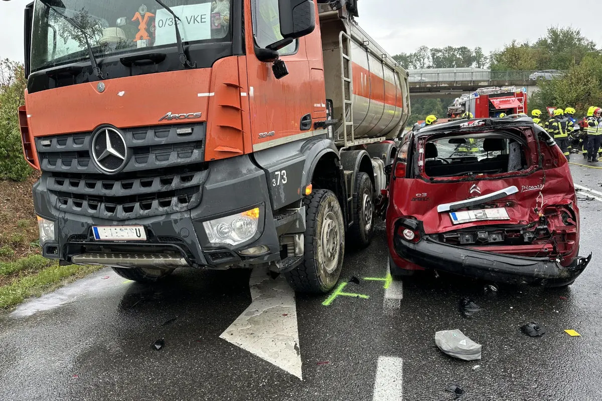 Ein roter LKW Mercedes kollidiert mit einem roten Citroën auf einer regennassen Straße. Feuerwehrleute sind im Hintergrund sichtbar.