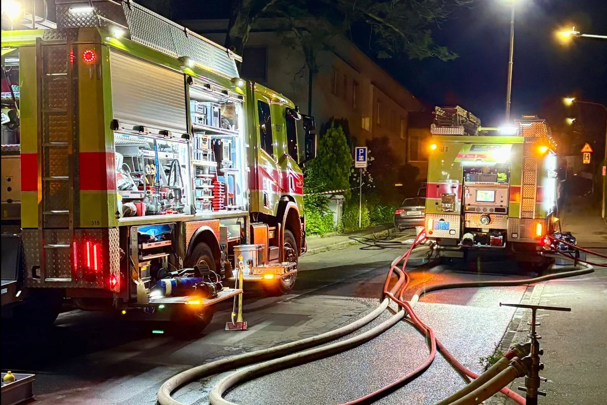 Zwei Feuerwehrfahrzeuge stehen bei Nacht auf einer Straße, umgeben von Schläuchen. Straßenlaternen beleuchten die Szene und ein Hydrant ist sichtbar.