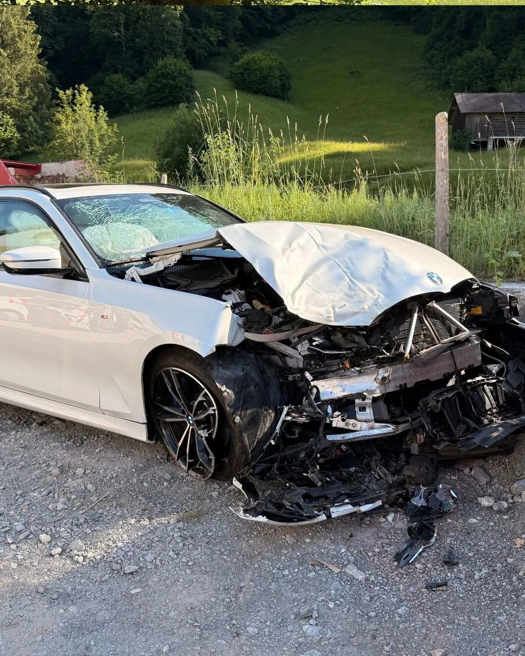 Ein weißer BMW mit stark beschädigter Front steht auf einer Kiesstraße. Im Hintergrund ist eine grüne Landschaft.